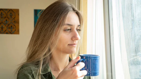 Depressed woman drinking coffee by the window at home. Thoughtful woman questioning life. Young adult girl watching outside