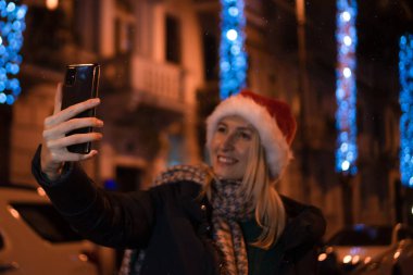 Noel Baba şapkalı mutlu genç kadın gece şehrinin Noel ışıklarının arka planında selfie çekiyor. Tatil ve insan kavramı. Telefonla eline odaklan.