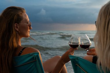 Mayolu, güneş gözlüklü, plajdaki katlanır sandalyelerde oturan iki genç kadın gün batımında kırmızı şarapla kadeh tokuşturuyor, arkadan fotoğraf çekiliyor.