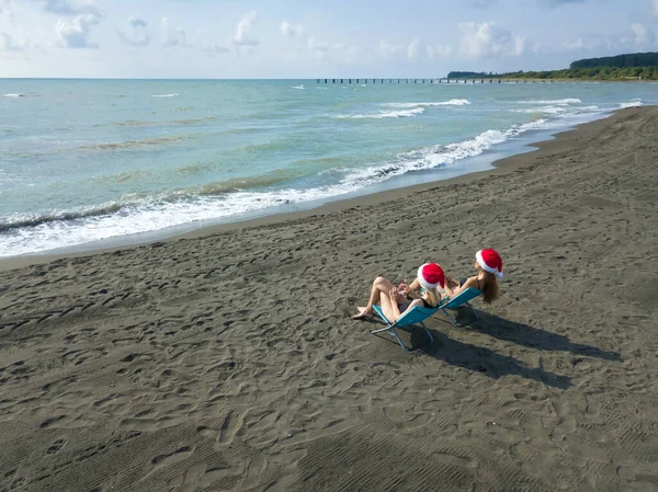 Mayolu ve Noel Baba şapkalı iki genç kadın gün batımından önce Karadeniz kıyısında bir bardak şarapla sandalyede oturuyorlar.