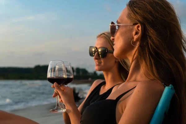 stock image Two young beautiful women in swimsuits, sunglasses are sitting on folding chairs on the beach with glasses of red wine at sunset. Focus on the brown-haired woman