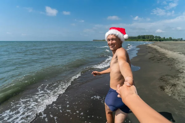 Happy smiling man in swimming trunks, Santa Claus hat holds a woman\'s hand and runs along the seashore on a sunny day. Come with me. Christmas holidays in a warm country