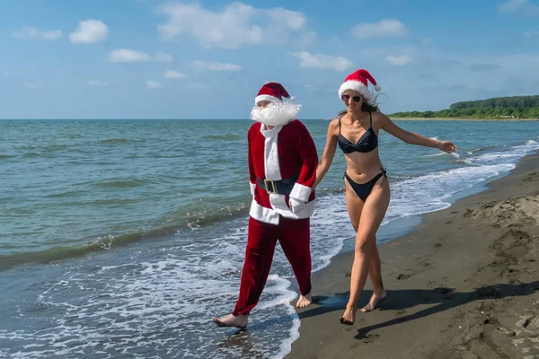 Święty Mikołaj Idzie Rękę Młodą Piękną Kobietą Bikini Kapeluszu Mikołaja — Zdjęcie stockowe
