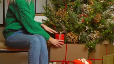 Yeşil kazaklı güzel beyaz kadın Noel ağacının altında hediyesini alır, açar ve mutlu bir şekilde gülümser. Ön planda köknar ağacı var. Ev Noel arifesi.