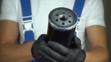 Mavi tulumlu araba tamircisi arabanın yağ filtresini elinde tutuyor ve siyah eldivenlerle döndürüyor. - Evet. Arabadaki motor ve otomobil parçalarındaki yağ değişimi kavramı. Sınıf işaretini göster.