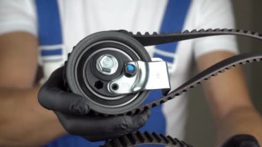 Mavi tulumlu oto tamircisi elinde siyah eldivenlerle gaz dağıtım mekanizmasının motor ve kuşak gerilimli kayış kayışlarıyla tutuyor. Arabada motorun kemerini değiştirme fikri. Sınıf işaretini göster