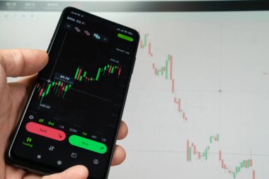 Şifreli para birimlerinin finansal piyasasını analiz etmek için analitik uygulamayı kullanarak akıllı bir telefonla bir adamın eline yakın çekim yapmak, bir monitörün arka planındaki yatırım artışı grafiği.