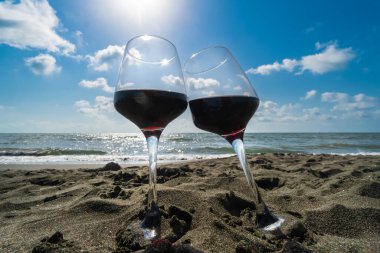 Sahilde kumsalda duran iki kadeh kırmızı şarap bulutlu ve güneş ışınlı mavi gökyüzüne karşı.