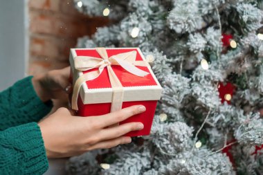 Yeşil kazaklı kadın elleri Noel ağacının yanında kırmızı bir Noel hediyesi kutusu tutuyor. Noel ve Mutlu Yıllar kavramı