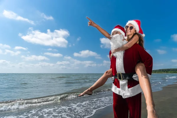 幸せなサンタクロースは 晴れた日に海辺の側に指差し指でサンタの帽子の笑顔の若い女性の背中に保持しています 暖かい国のクリスマス休暇を祝う — ストック写真