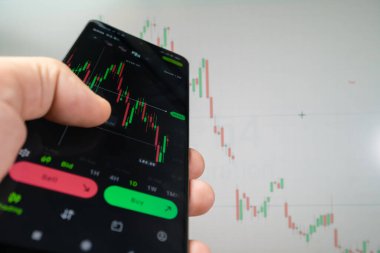 Şifreli para birimlerinin finansal piyasasını analiz etmek için analitik uygulamayı kullanarak akıllı bir telefonla bir adamın eline yakın çekim yapmak, bir monitörün arka planındaki yatırım artışı grafiği.