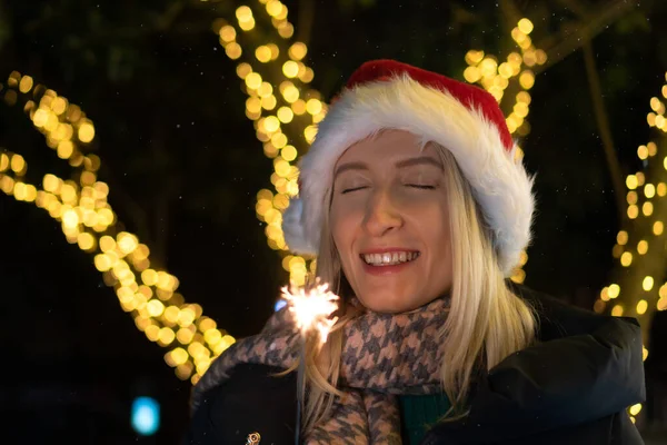 stock image Happy smiling woman in a Santa Claus hat with her eyes closed holds sparklers and makes a wish on the street against the background of lights at night