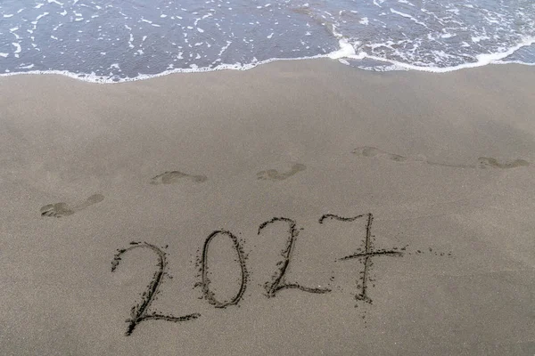 Küçük bir dalga ve insan ayağı izleriyle dolu kumlu bir sahilde mutlu yıllar 2027 mesajı at. Yeni Yıl 2027 'nin soyut arkaplan fotoğrafı