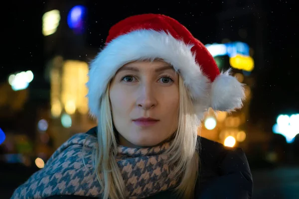 stock image Portrait of a young serious woman in a santa claus hat, looking into the camera, on the street of the night city