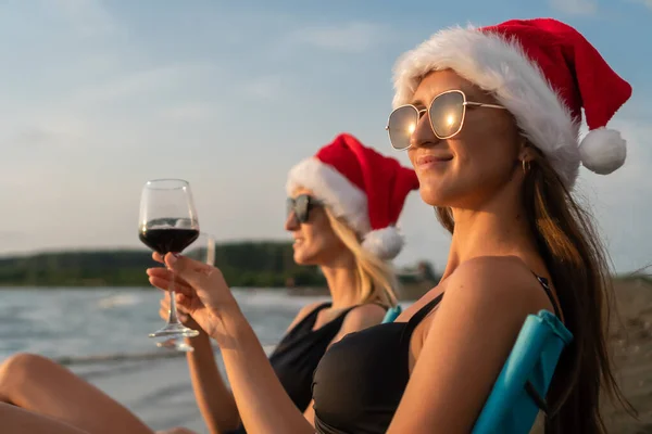 Two young beautiful slender women in swimsuits, sunglasses, Santa Claus hats are sitting on folding chairs on the sandy beach with glasses of red wine at sunset. Focus on the brown-haired woman