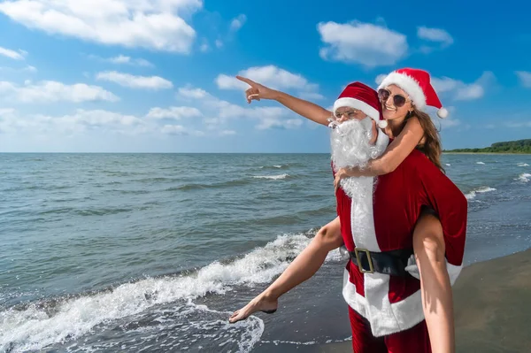 幸せなサンタクロースは 晴れた日に海辺の側に指差し指でサンタの帽子の笑顔の若い女性の背中に保持しています 暖かい国のクリスマス休暇を祝う — ストック写真