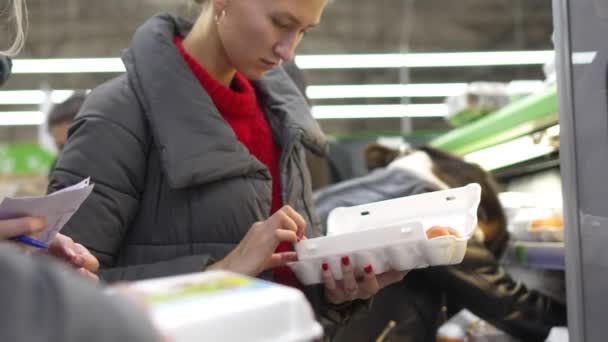 一个漂亮的女人在超市里买鸡蛋 打开包装 检查里面的东西是否完整在购买前检查货物 消费者篮子 很多有背景的人 — 图库视频影像