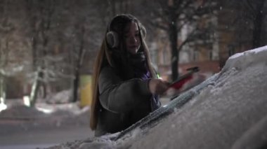 Young girl in headphones or hat listens to music and removes snow with brush from car at night or in evening against background of falling snow in winter and night city with lights. Fabulous weather