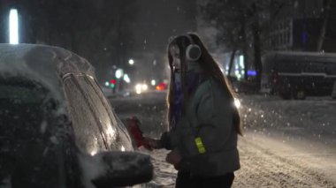 Young girl in headphones or hat listens to music and removes snow with brush from car at night or in evening against background of falling snow in winter and night city with lights. Fabulous weather