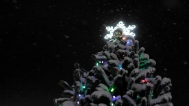 Christmas tree view of bottom against background of falling snow and black night sky. Christmas Eve and New Year's Eve, fir tree with glowing snowflake on top. Fabulous atmosphere.