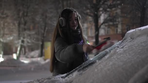 ヘッドフォンや帽子の若い女の子は音楽に耳を傾け 夜や夜に車からブラシで雪を削除します 素晴らしい天気 — ストック動画