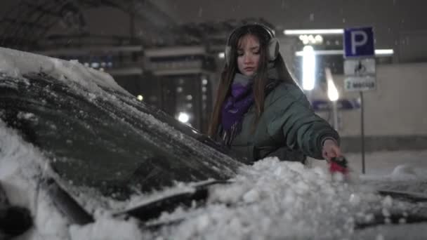 ヘッドフォンや帽子の若い女の子は音楽に耳を傾け 夜や夜に車からブラシで雪を削除します 素晴らしい天気 — ストック動画