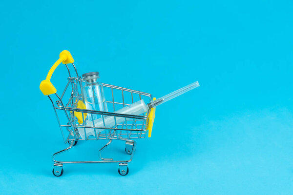 Shopping cart with a medical ampoule and a syringe on a blue background, copy the space. Concept of purchasing medicines, increasing drug prices