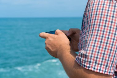 Close-up of a man in a plaid shirt sitting on the seashore on a sunny day and holding a mobile phone in his hands