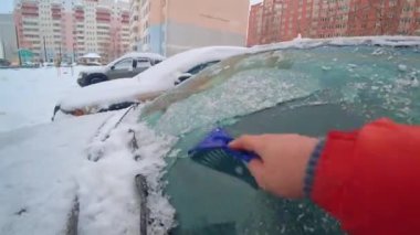 Birinci kişi buz kazıyıcı ile arabadaki buzları temizler. Buz kazıyıcıya odaklan. Soğuk, karlı ve soğuk bir sabah. Aksiyon Kamerası Canlı Kamera. Şehrin arka planına ve sık binalara karşı.