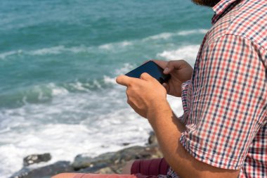 Man in a plaid shirt sitting on the seashore on a sunny day and holding a mobile phone in his hands