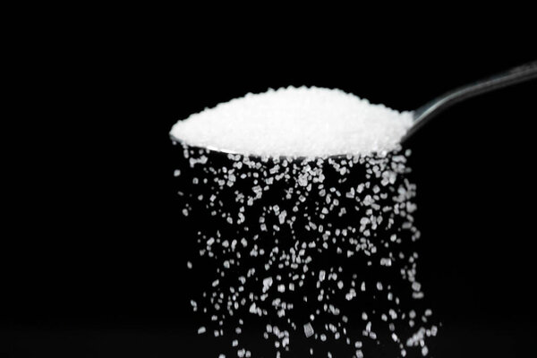 Macro photography of a teaspoon with white sugar on a black background, copy space. White sugar is poured from a teaspoon.