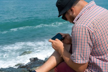 Man in a plaid shirt sitting on the seashore on a sunny day and holding a mobile phone in his hands