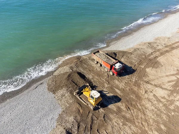 Drone view of a bulldozer and a truck with small stones on the seashore on a sunny day. Concept of strengthening the embankment, beach of the sea.
