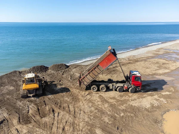 View from a drone on a bulldozer and a dump truck that dropped stones on the sea embankment on a sunny day. Concept of strengthening the coastal line of the beach.