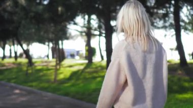 Günışığında şehir parkında yürüyen çağdaş giyinmiş genç güzel bir kadın. Kamera hareket eder, saçlar rüzgarda gelişir. Şehir parkı yaz akşamları ya da sabahları. Arka görünüm.