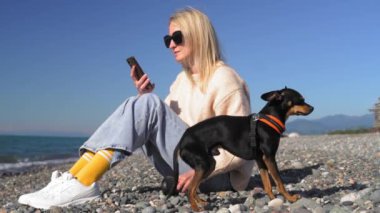 Küçük oyuncak teriyer köpeği, deniz manzarasına karşı tasmalı, elinde akıllı telefon olan bir kadının yanında. Neşe yürüyüşü ve kumsal kokusu, sıcak güneş ve hafif deniz esintisi hissi. Deniz kenarında iyi yürüyüşler..