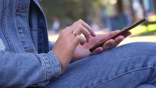 公園に座っているジーンズのデニムジャケットの女性は 彼女のスマートフォンを吸収した 彼女の指は 仮想世界に吸収されたコンテンツをスクロールするときに画面をタップします 自然に囲まれたエンターテイメントは遠くに見えます — ストック動画