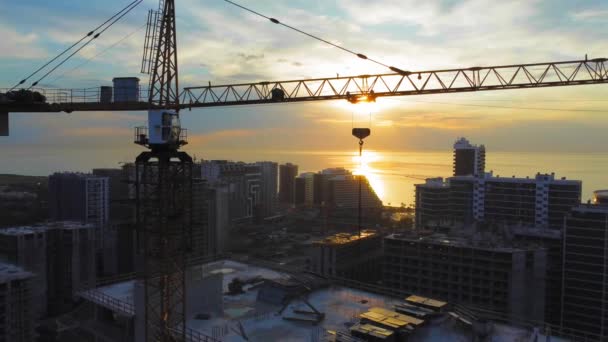 海や海の上の太陽を背景に建設クレーン付きの新しい近代的なアパートの建物 高層ビル 日没時に市内のクレーン建設現場のドローンビュー 請負業者 — ストック動画