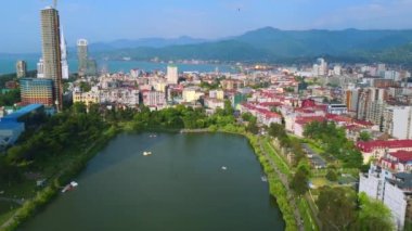 İnsansız hava aracı, 6 Mayıs 'ta çocuklar için parkın yanı sıra güneşli bir günde deniz kenarındaki çocuk parkında gökdelenlerin ve dağların zeminine karşı gölle parkta uçuyor. Gürcistan 'ın Batumi kentine bakın