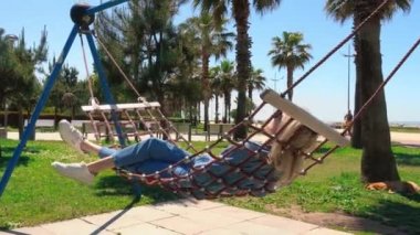 Güneşli bir günde şehir parkında hamakta dinlenen bir kadın şehir ve palmiye ağaçlarına karşı. Seyahat ve rahatlatıcı yaşam tarzı kavramı. Boş zaman ve rahatlama açık havada tek başına.
