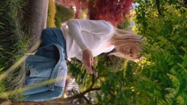 Güzel ve güzel bir genç kız, güneşli bir günde yeşil parkta otururken telefon kullanıyor ve dinleniyor. Gülümsüyor ve telfona bakıyor. Sarışın. Dikey video