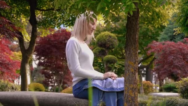 電話とリラックスを使用して魅力的でかなり若い女の子は 晴れた暖かい日に美しい緑の公園のベンチに座っています 彼女は笑顔で電話を調べている ブロンド — ストック動画