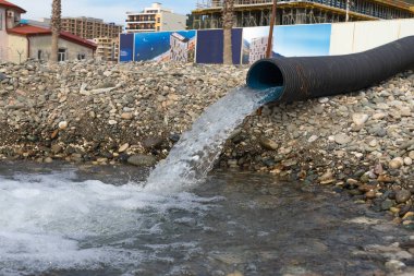 Büyük plastik bir su borusunun yakın plan çekimi. Büyük bir su akıntısı denizdeki çakıl taşı plajına taşar.
