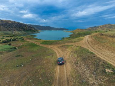 Düşük dağların arasında göle ya da su deposuna yaklaşan bir arabanın drone görüntüsü. İnsansız hava aracından güzel bir manzara. Dinlenme kavramı, seyahat.