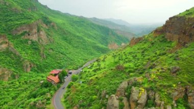 Yazın günbatımında yeşil çayırlardaki yol manzarası. Kırsal yol, dağlar, orman yukarıdan insansız hava aracı görüntüsü. Yol, güneş ışınları, ağaçlar, tepeler, yeşil çimenler, bulutlar olan güzel bir manzara. Ermenistan