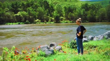 Kot pantolonlu ve tişörtlü genç bir kadın dağların arka planında bir dağ nehrinin yanında duruyor. Nehir, ağaçlar, gelincikler ve arkadan bir kız. Seyahat kavramı, yaşam tarzı.