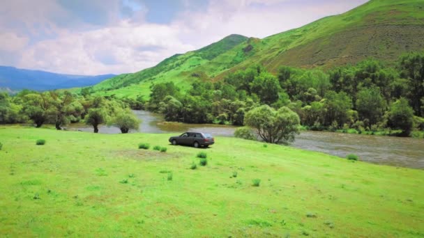 山と緑の牧草地を背景に山の川の銀行に沿って車に乗る 旅行のコンセプト 日当たりの良い緑の牧草地のドローンビュー 田舎を走る 美しい魔法の景色 — ストック動画