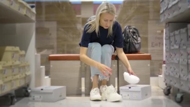漂亮的金发姑娘坐在时尚精品店里试鞋 她选择了白色的运动鞋 围绕着许多有风格的盒子和架子 买鞋子 — 图库视频影像