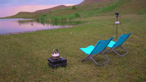 水壶在湖边的营地 夏天在湖边的野营炉上煮咖啡 美丽的晴天云和蓝天 在旅行时煮咖啡 附近有野营用的椅子 — 图库视频影像