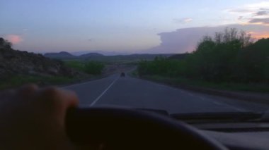 Arabayla gezmek, akşam otobanda seyahat etmek. Ön pencereden doğaya, gün batımına bak. Güneşin bulutlarında güzel turuncu-kırmızı gökyüzü. Sürücünün birinci şahıs görüntüsü, yolda direksiyon..